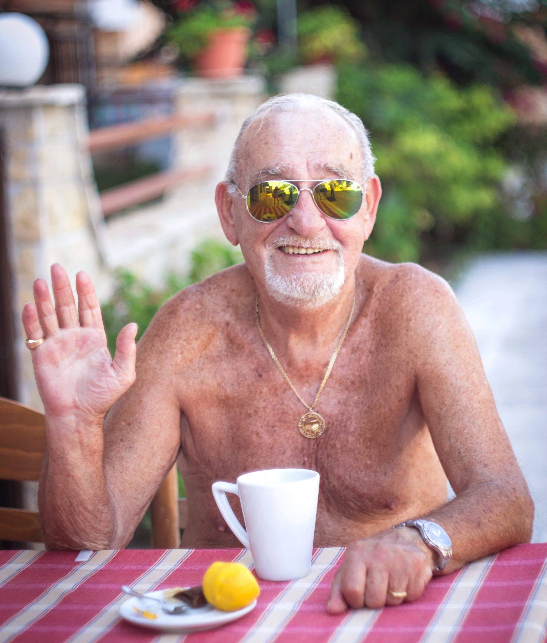 Friendly older gentleman sitting shirtless at a table smiling and waving